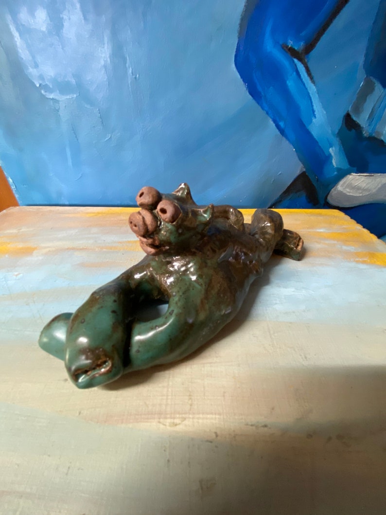 May include: A handcrafted ceramic sculpture in the shape of a hand, with a creature-like head. The sculpture is glazed in shades of green and brown. The hand has a hole in the center. The head has three eyes and a snout.
