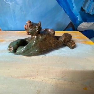 May include: A handmade ceramic sculpture of a reclining cat in shades of green and brown. The cat is lying on its back with its paws outstretched. The sculpture has a glossy finish and is set against a blue and yellow background.