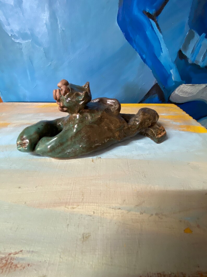 May include: A handmade ceramic sculpture of a reclining cat in shades of green and brown. The cat is lying on its back with its paws outstretched. The sculpture has a glossy finish and is set against a blue and yellow background.