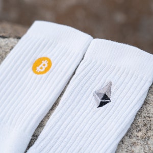 Könnte beinhalten: Zwei weiße Socken mit gestickten Logos. Eine Socke hat ein gelbes Bitcoin-Symbol, die andere ein graues Ethereum-Symbol.