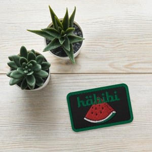 May include: A black and green embroidered patch with the word "Habibi" and a red watermelon slice on a white wooden surface with two green succulents in white pots.