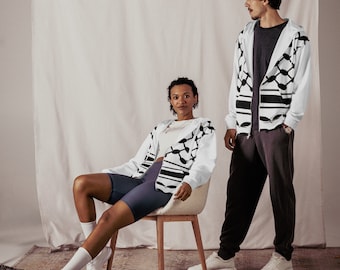 May include: Two people wearing white and black patterned jackets. The person on the left is sitting on a chair and wearing black shorts. The person on the right is standing and wearing grey pants.