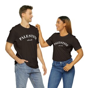 Peut inclure: Deux personnes portant des t-shirts noirs avec le mot "PALESTINE" en lettres blanches et le mot arabe pour Palestine en dessous. Les t-shirts sont de conception simple avec un col rond et des manches courtes.