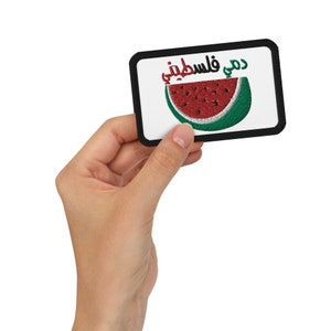 May include: Embroidered patch featuring a watermelon slice design with the text in Arabic. The watermelon has red and green colors, and the patch has a black border. The patch is rectangular and held by a hand.