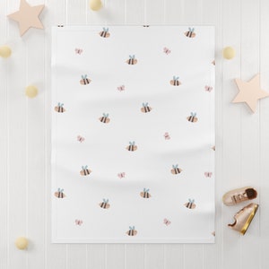 May include: White baby blanket with a repeating pattern of bees and butterflies. The bees are yellow and brown with black stripes and blue wings. The butterflies are pink.