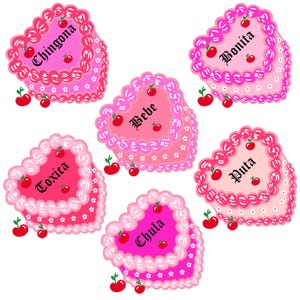 May include: Seven pink heart-shaped cakes with white frosting and pink and white sprinkles. Each cake has a different Spanish word written in black on top: Chingona, Bonita, Bebe, Toxica, Puta, Chula.