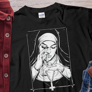 Könnte beinhalten: Schwarzweiß-Illustration einer Nonne mit weißem Habit und Rosenkranz, die einen zusammengerollten Geldschein und eine Tüte mit weißem Pulver hält. Die Illustration befindet sich auf einem schwarzen T-Shirt.
