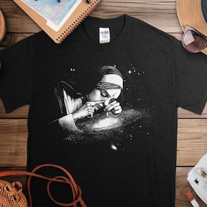 May include: A black t-shirt with a white graphic of a person looking through a magnifying glass at a galaxy. The person is wearing a hat and has a serious expression.