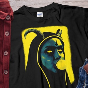 Puede incluir: Camiseta negra con un gráfico amarillo y negro de una monja con cuernos, una cruz y una burbuja. La monja tiene la piel azul y los ojos blancos.
