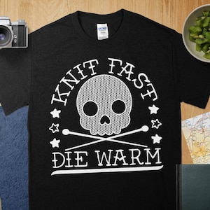 May include: Black t-shirt with a white graphic featuring a skull made of a knitted pattern, crossed knitting needles, and the text "KNIT FAST DIE WARM". The design is surrounded by stars.