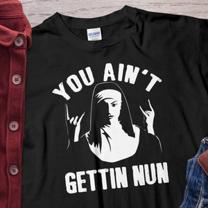 Puede incluir: Camiseta negra con un gráfico blanco de una monja haciendo un gesto de rock and roll con la mano. El texto "YOU AIN'T GETTIN NUN" está impreso encima y debajo de la ilustración de la monja. La camiseta se muestra sobre una superficie de madera.
