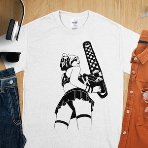 Könnte beinhalten: Weißes T-Shirt mit einer schwarz-weißen Grafik einer Person, die eine Kettensäge hält. Die Person trägt einen Rock und ein Geschirr. Das T-Shirt wird auf einer Holzoberfläche mit anderen Gegenständen präsentiert.