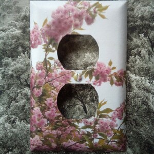 以下が含まれることがあります： ピンクの桜の花が咲いた花柄の白いコンセントカバー。カバーには標準的なコンセントが2つあります。