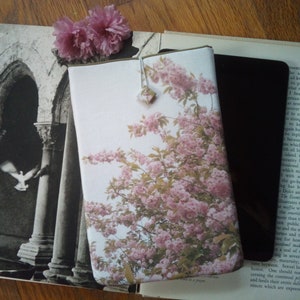 May include: A white book cover with a floral design featuring pink blossoms and green leaves. The cover is slightly textured and has a subtle sheen. The book is lying on a wooden surface.