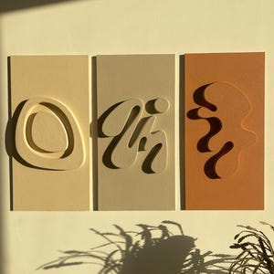 Może przedstawiać: Trzy prostokątne dekoracje ścienne z wyciętymi abstrakcyjnymi kształtami. Lewa część jest beżowa z jasnobrązowym kształtem, środkowa część jest jasnobrązowa z ciemnobrązowym kształtem, a prawa część jest pomarańczowa z ciemnopomarańczowym kształtem.