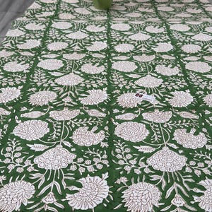 Puede incluir: Un mantel con estampado floral verde y blanco. El dise&ntilde;o presenta grandes flores y hojas blancas sobre un fondo verde oscuro. El mantel es rectangular y parece estar hecho de tela.