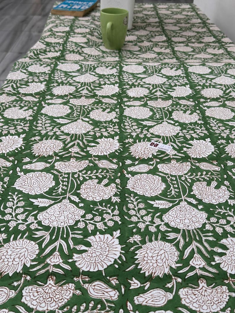 Puede incluir: Un mantel con estampado floral verde y blanco. El dise&ntilde;o presenta grandes flores y hojas blancas sobre un fondo verde oscuro. El mantel es rectangular y parece estar hecho de tela.