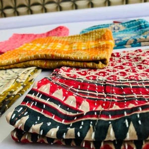 May include: A collection of folded fabric pieces featuring intricate white running stitch embroidery. The textiles display various patterns and colors, including red, black, yellow, orange, and blue, with geometric and floral motifs visible throughout.