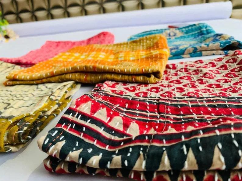 May include: A collection of folded fabric pieces featuring intricate white running stitch embroidery. The textiles display various patterns and colors, including red, black, yellow, orange, and blue, with geometric and floral motifs visible throughout.