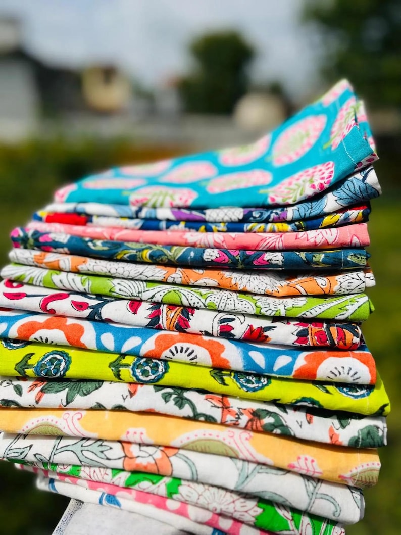 K&ouml;nnte beinhalten: Ein Stapel bunter Stofft&uuml;cher mit verschiedenen floralen und abstrakten Mustern. Die T&uuml;cher sind in den Farbt&ouml;nen Blau, Rosa, Gr&uuml;n, Gelb und Wei&szlig; gehalten. Das Bild wurde im Freien mit unscharfem Hintergrund aufgenommen.