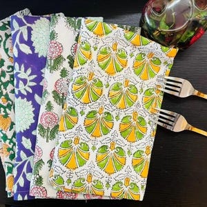 K&ouml;nnte beinhalten: Ein Stapel bunter Stoffservietten mit floralen Mustern in Orange, Lila und Gr&uuml;n. Die Servietten sind gefaltet und auf einer dunklen Oberfl&auml;che angeordnet, mit zwei Gabeln und einer dekorativen Glasschale.