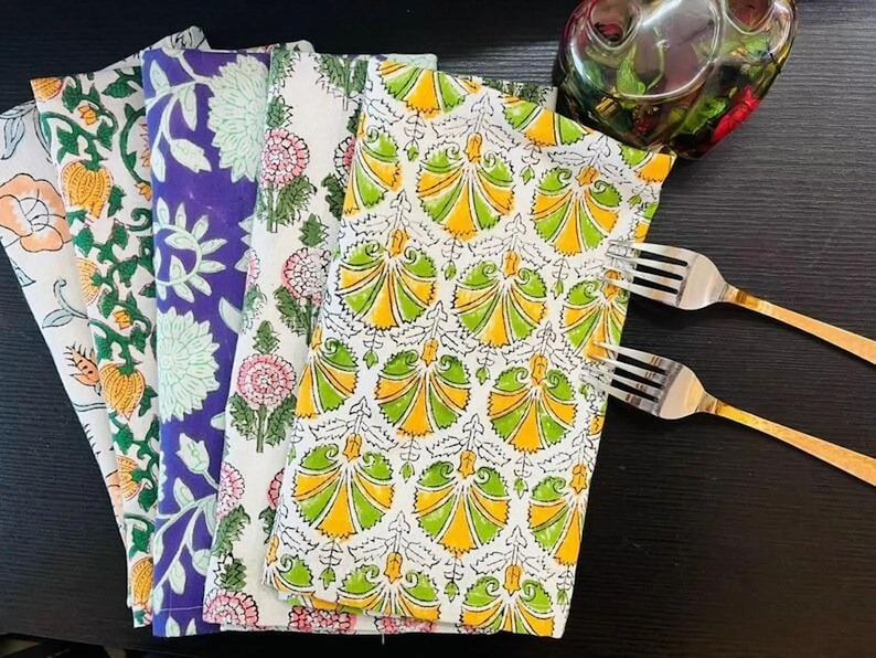 K&ouml;nnte beinhalten: Ein Stapel bunter Stoffservietten mit floralen Mustern in Orange, Lila und Gr&uuml;n. Die Servietten sind gefaltet und auf einer dunklen Oberfl&auml;che angeordnet, mit zwei Gabeln und einer dekorativen Glasschale.