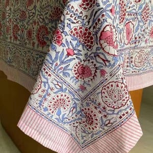 Pode incluir: Uma toalha de mesa branca com um padrão floral em tons de rosa, bordô e azul. O design apresenta grandes flores e folhas, com uma borda listrada em rosa. O tecido está drapeado sobre uma mesa de madeira.