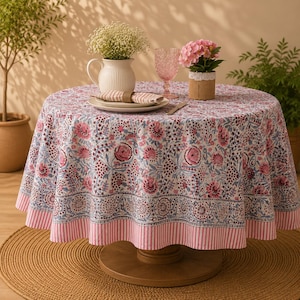 May include: A round table covered with a white tablecloth featuring a pink and blue floral pattern and a pink striped border. On the table sits a white pitcher with baby's breath, a pink glass, a small potted pink flower, and a folded napkin.