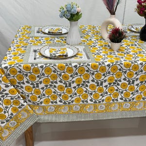 Op de afbeelding: Een witte tafelkleed met een geel bloemenpatroon, gedekt met bijpassende placemats, borden en servetten. De tafel is versierd met vazen met bloemen. Het tafelkleed heeft een rand met een streepjesmotief.