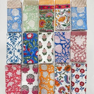 K&ouml;nnte beinhalten: Eine Sammlung gefalteter Stoffservietten mit verschiedenen Mustern. Die Servietten zeigen florale und geometrische Designs in Farben wie Blau, Gr&uuml;n, Rot und Orange. Einige haben gestreifte R&auml;nder. Die Servietten sind ordentlich auf einer wei&szlig;en Oberfl&auml;che angeordnet.
