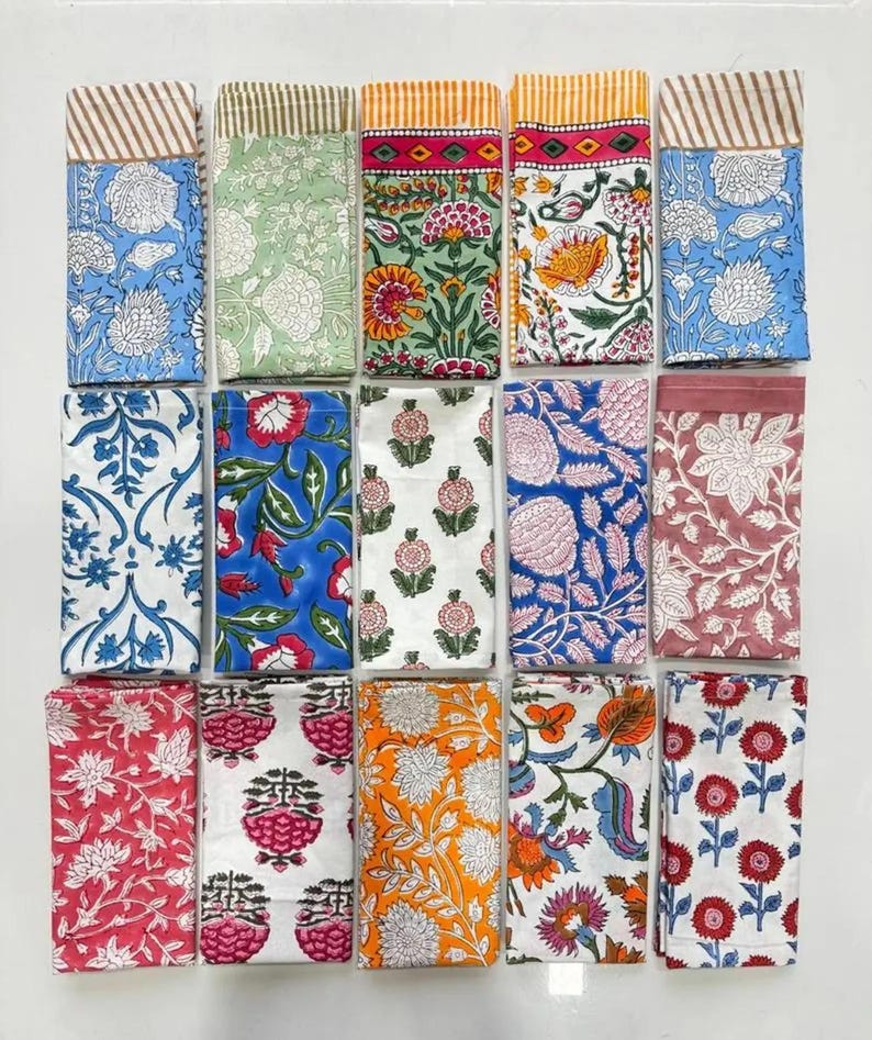 K&ouml;nnte beinhalten: Eine Sammlung gefalteter Stoffservietten mit verschiedenen Mustern. Die Servietten zeigen florale und geometrische Designs in Farben wie Blau, Gr&uuml;n, Rot und Orange. Einige haben gestreifte R&auml;nder. Die Servietten sind ordentlich auf einer wei&szlig;en Oberfl&auml;che angeordnet.