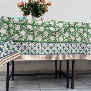 Puede incluir: Mantel verde floral con un patr&oacute;n de flores blancas. El borde presenta un dise&ntilde;o de abanico azul y blanco con rayas marrones. Un jarr&oacute;n de flores est&aacute; sobre la mesa.