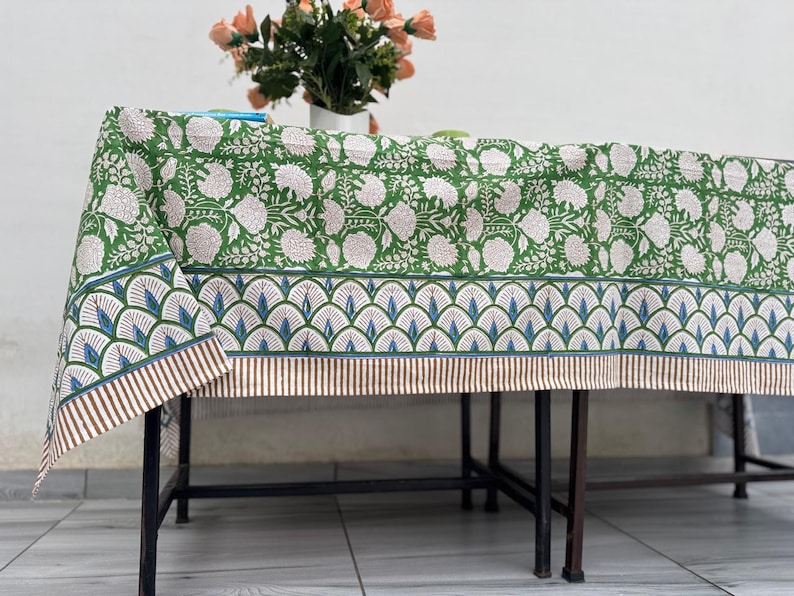 Puede incluir: Mantel verde floral con un patr&oacute;n de flores blancas. El borde presenta un dise&ntilde;o de abanico azul y blanco con rayas marrones. Un jarr&oacute;n de flores est&aacute; sobre la mesa.