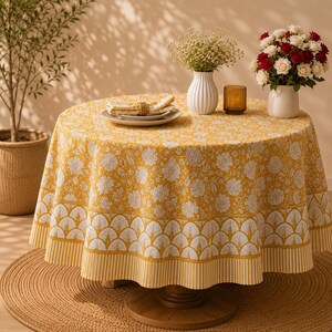 Pu&ograve; includere: Un tavolo rotondo coperto da una tovaglia giallo senape con un motivo floreale bianco e un bordo smerlato. Sul tavolo si trovano un piatto bianco con un tovagliolo piegato, un vaso bianco con piccoli fiori bianchi e un portacandele in vetro.