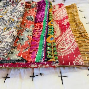 May include: A stack of five folded fabric textiles featuring diverse patterns including floral, paisley, and striped designs in colors of cream, red, magenta, green, yellow, and brown, all finished with visible white running stitch embroidery.
