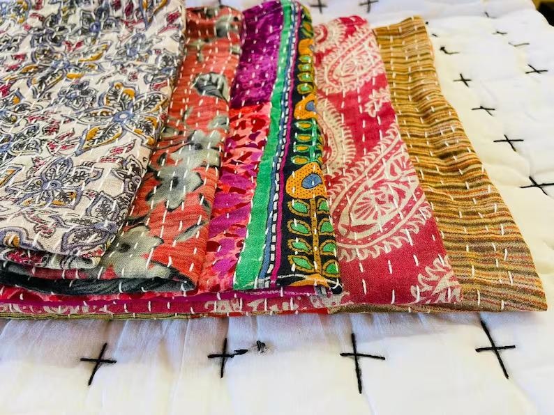 May include: A stack of five folded fabric textiles featuring diverse patterns including floral, paisley, and striped designs in colors of cream, red, magenta, green, yellow, and brown, all finished with visible white running stitch embroidery.