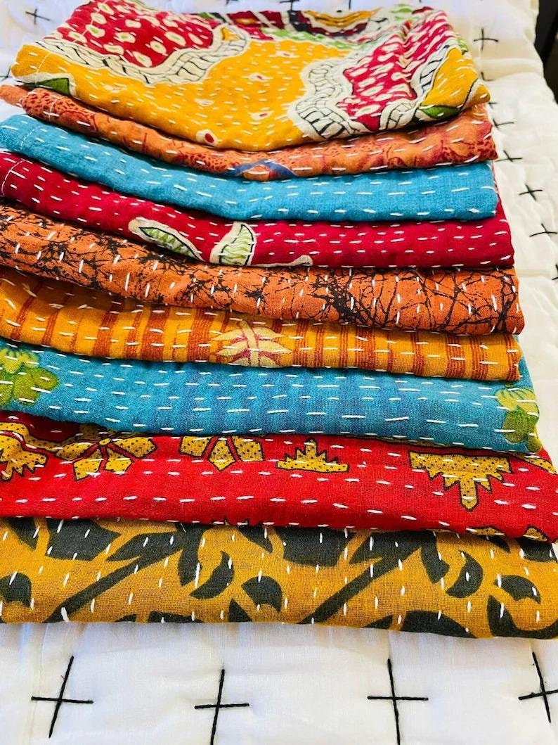 May include: A stack of folded fabric quilts featuring various patterns and colors including yellow, orange, blue, red, and black. Each layer displays intricate white running stitch details across the surface, creating a textured appearance.