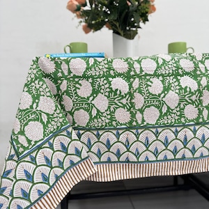 Puede incluir: Un mantel verde con un estampado floral blanco y un borde decorativo. El borde presenta un dise&ntilde;o repetitivo de elementos azules y marrones. El mantel est&aacute; sobre una mesa, con un jarr&oacute;n de flores color melocot&oacute;n en el fondo.