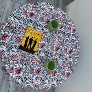 Pu&ograve; includere: Una tovaglia rotonda con un motivo floreale ripetuto nei toni del rosa, blu e grigio scuro su sfondo bianco. Un libro intitolato The 3 Mistakes of My Life e due tazze in ceramica verde chiaro sono posizionati sulla superficie del tessuto.