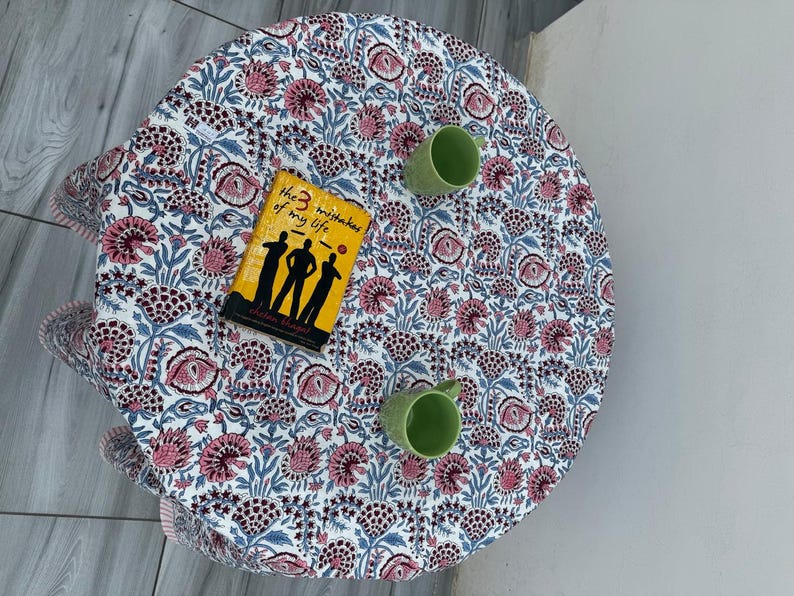 Pu&ograve; includere: Una tovaglia rotonda con un motivo floreale ripetuto nei toni del rosa, blu e grigio scuro su sfondo bianco. Un libro intitolato The 3 Mistakes of My Life e due tazze in ceramica verde chiaro sono posizionati sulla superficie del tessuto.