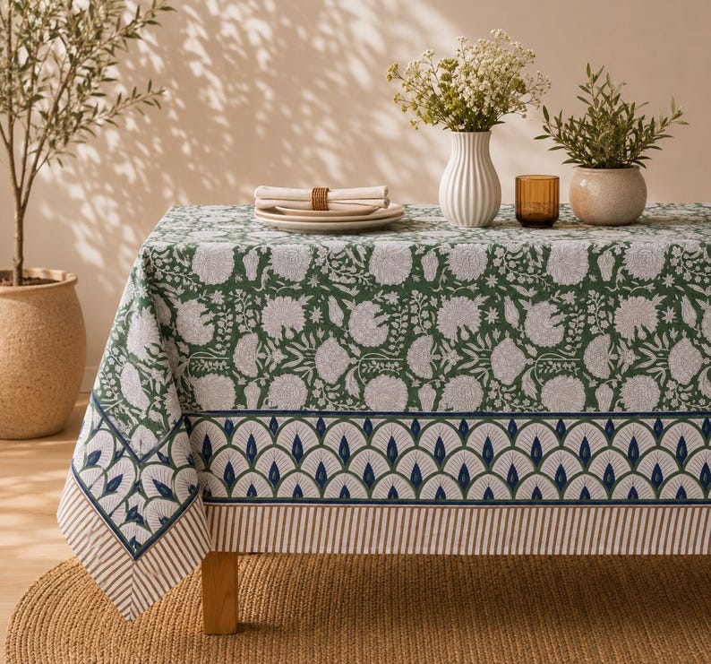 Pu&ograve; includere: Una tovaglia rettangolare caratterizzata da un motivo floreale verde e bianco sulla superficie superiore, bordata da un design smerlato blu e bianco e da un bordo a righe sottili. Il tessuto copre un tavolo in legno, mostrando una stampa botanica.