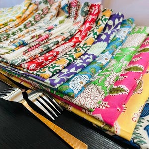 K&ouml;nnte beinhalten: Ein Stapel bunter, gemusterter Stoffservietten in verschiedenen Farben, darunter Rot, Gr&uuml;n, Lila und Blau. Die Servietten weisen florale Designs auf und sind neben zwei silbernen und goldenen Gabeln angeordnet.
