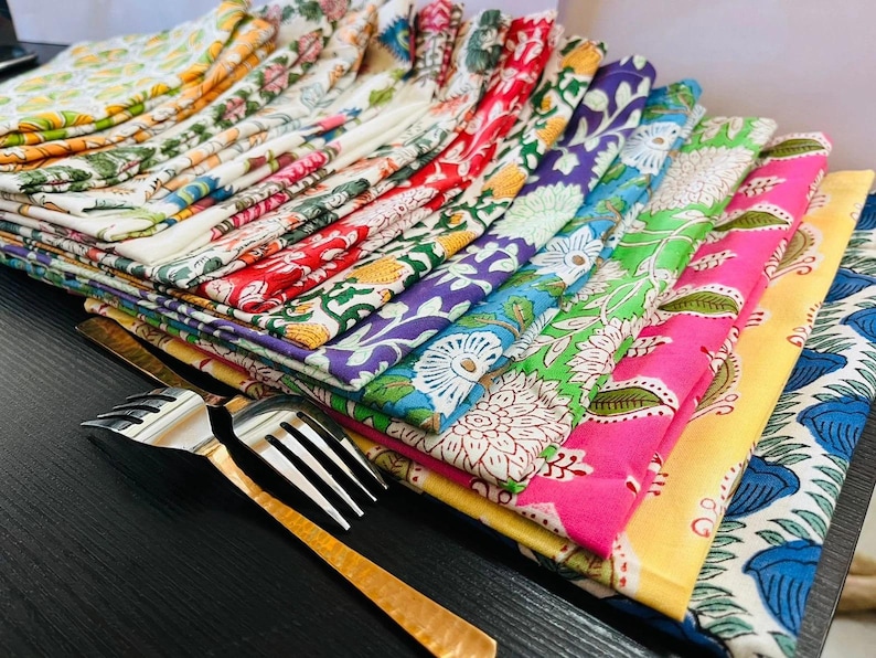 K&ouml;nnte beinhalten: Ein Stapel bunter, gemusterter Stoffservietten in verschiedenen Farben, darunter Rot, Gr&uuml;n, Lila und Blau. Die Servietten weisen florale Designs auf und sind neben zwei silbernen und goldenen Gabeln angeordnet.