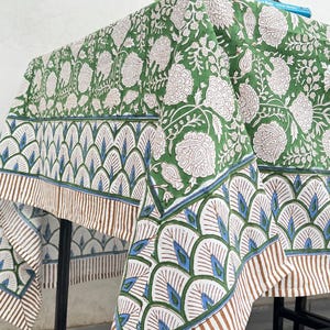 Puede incluir: Un mantel con estampado floral verde y blanco con un borde decorativo. El borde presenta un patr&oacute;n repetitivo de formas de abanico blancas, azules y marrones. El mantel est&aacute; drapeado sobre una mesa de metal negro.