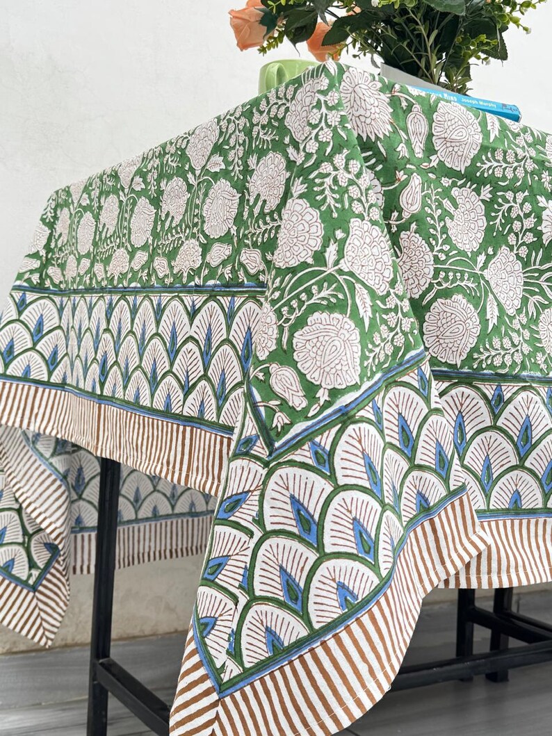 Puede incluir: Un mantel con estampado floral verde y blanco con un borde decorativo. El borde presenta un patr&oacute;n repetitivo de formas de abanico blancas, azules y marrones. El mantel est&aacute; drapeado sobre una mesa de metal negro.