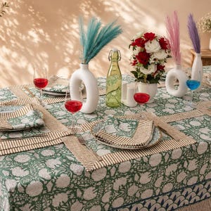 Pu&ograve; includere: Un tavolo da pranzo con una tovaglia a motivo floreale verde e bianco e tovagliette abbinate. Il tavolo presenta vasi in ceramica bianca con erba della pampa colorata, una bottiglia di vetro verde, piatti con tovaglioli e calici.