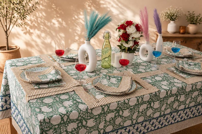 Pu&ograve; includere: Un tavolo da pranzo con una tovaglia a motivo floreale verde e bianco e tovagliette abbinate. Il tavolo presenta vasi in ceramica bianca con erba della pampa colorata, una bottiglia di vetro verde, piatti con tovaglioli e calici.