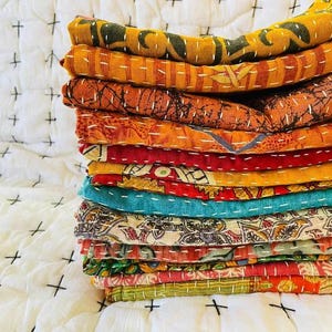 Puede incluir: Una pila de coloridas colchas kantha estampadas. Las colchas presentan varios colores, incluyendo naranja, rojo y turquesa, con costuras blancas. Las colchas dobladas están apiladas, creando una exhibición visualmente atractiva.