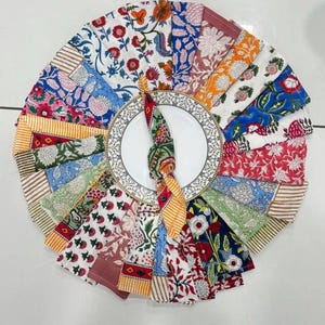 K&ouml;nnte beinhalten: Eine kreisf&ouml;rmige Anordnung farbenfroher Stoffservietten mit floralen Mustern um einen wei&szlig;en Teller mit dekorativem Rand. Die Servietten zeigen verschiedene Designs in Rot-, Blau-, Orange- und Gr&uuml;nt&ouml;nen.