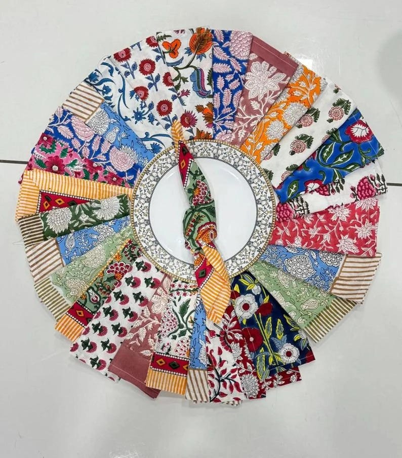 K&ouml;nnte beinhalten: Eine kreisf&ouml;rmige Anordnung farbenfroher Stoffservietten mit floralen Mustern um einen wei&szlig;en Teller mit dekorativem Rand. Die Servietten zeigen verschiedene Designs in Rot-, Blau-, Orange- und Gr&uuml;nt&ouml;nen.
